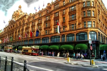 Exterior of Harrods in London, showcasing its iconic architecture with ornate details, green awnings, and flags of various countries, with pedestrians and double-decker buses passing by.