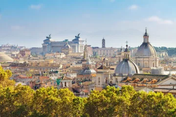 Panoramic view across the rooftops and domes of Rome under a bright blue sky.
