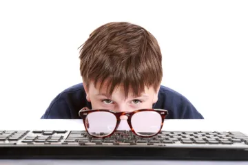 A boy peering over a keyboard with a pair of glasses propped in front of him.