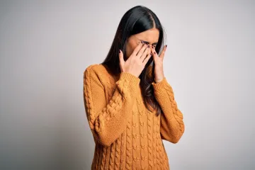 A woman in an orange jumper pressing both hands over her closed eyes.