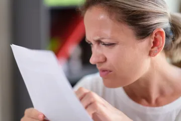 A woman frowning and squinting as she reads a sheet of paper held close to her face.