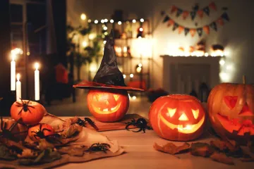 Three carved jack-o'-lanterns glowing on a wooden table — the central one wearing a black witch's hat — surrounded by lit candles, plastic spiders and autumn leaves, with string lights and bunting on shelves behind.