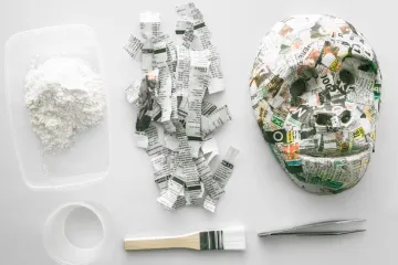 Flat lay of papier-mâché supplies on a white surface: a tub of flour paste, torn strips of newspaper, a finished newspaper mask, a water pot, a brush and tweezers.