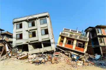 Two multi-storey concrete buildings left leaning and partly collapsed after an earthquake, surrounded by piles of brick and concrete rubble under a clear blue sky.