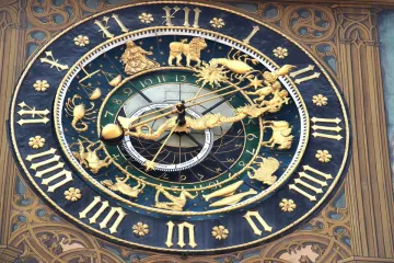 An ornate gilded astronomical clock on a painted Gothic façade, with a ring of zodiac signs and figures, black Gothic hour numerals, a golden sun-and-dragon hand and a month dial labelled in German.
