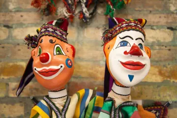 Two brightly painted papier-mâché puppet figures with clown faces, colourful woven hats and striped folk clothing, against a brick wall.