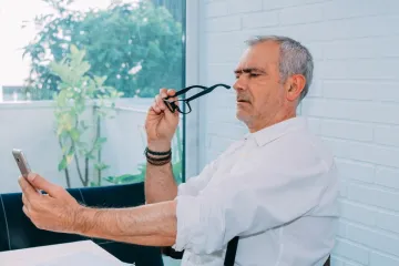 A man holding his glasses away from his face and a phone at arm's length to read the screen.