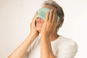 A man holding a folded warm cloth across his closed eyes with both hands.
