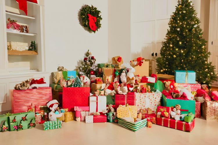 A festive room decorated for Christmas, featuring a large Christmas tree with lights and a variety of wrapped gifts in colorful paper arranged underneath. There are also several stuffed animals and toys among the presents. A wreath with a red bow hangs on the wall, and the scene includes a smaller decorated tree and shelves with additional holiday decorations.