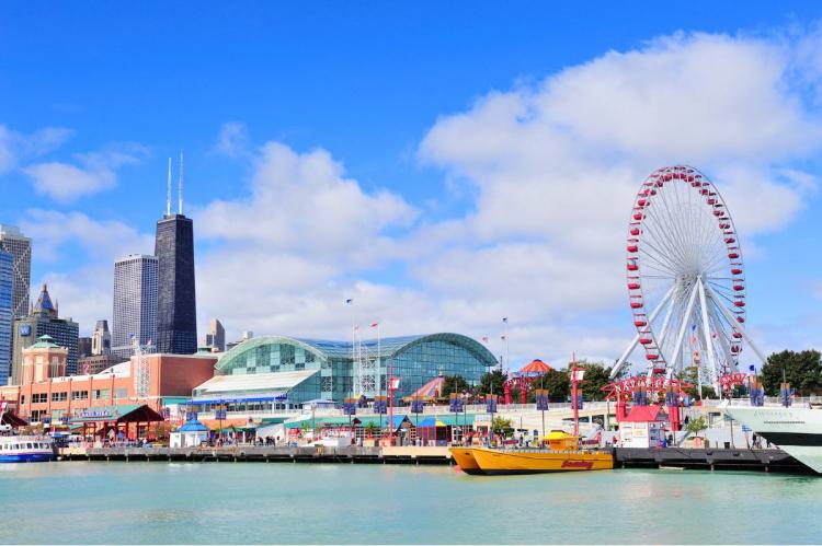 Navy Pier in Chicago with a large Ferris wheel, colorful buildings, and boats, with the John Hancock Center in the background.