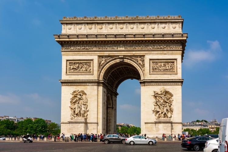 The Arc de Triomphe in Paris, a large stone monument with detailed carvings, surrounded by cars and visitors.