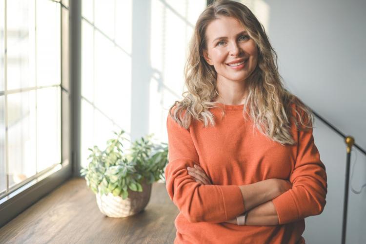 A smiling middle-aged woman with wavy blonde hair, wearing an orange sweater, stands by a window with natural light and a potted plant nearby.