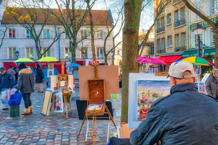 Street scene in Montmartre, Paris, with artists painting on easels in an open square, colorful umbrellas, and surrounding Parisian buildings under a tree-lined setting.