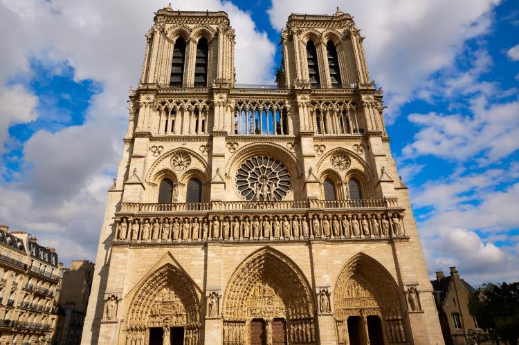 Front view of the Notre-Dame Cathedral in Paris with its twin towers, detailed facade, and central rose window under a partly cloudy blue sky.