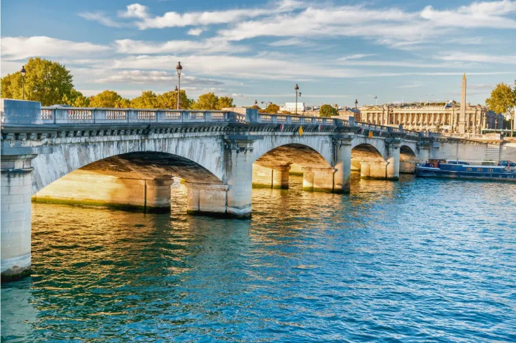 Sunlit stone bridge crossing the Seine River in Paris, with reflections on the water and historical buildings in the background.