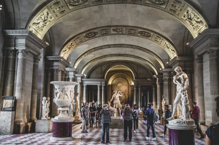 Interior of the Louvre Museum featuring classical statues, marble urns, and an arched ceiling.