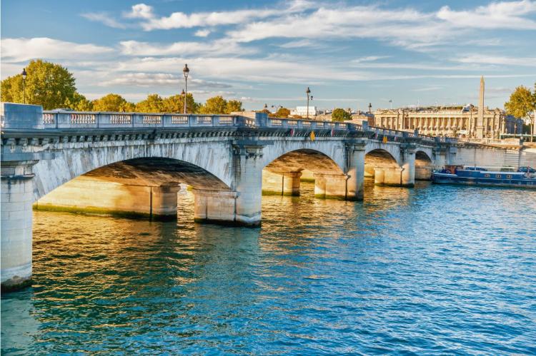 Sunlit stone bridge crossing the Seine River in Paris, with reflections on the water and historical buildings in the background.