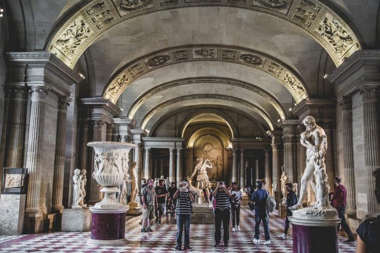 Interior of the Louvre Museum featuring classical statues, marble urns, and an arched ceiling.