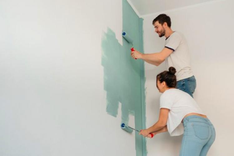 A man and woman using paint rollers to apply a soft green color to a white wall, painting together in a home improvement project.