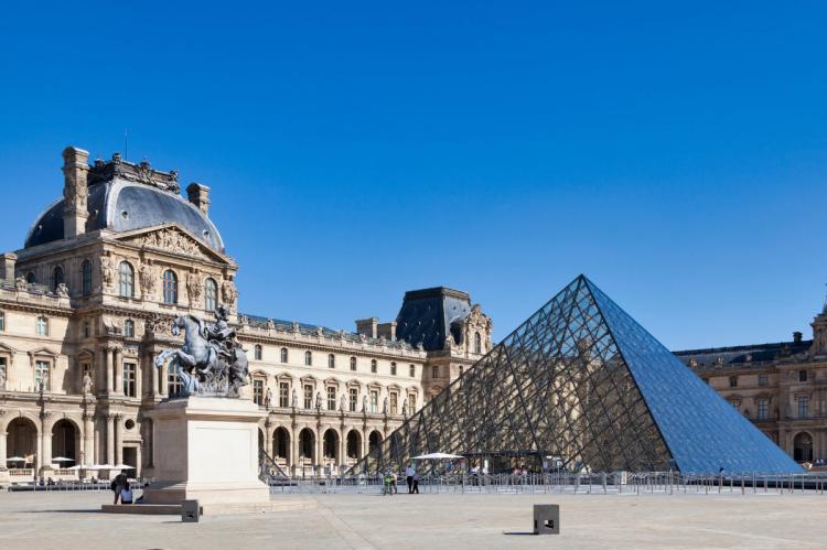 The glass pyramid of the Louvre Museum in Paris, set against the historic museum buildings and a clear blue sky.