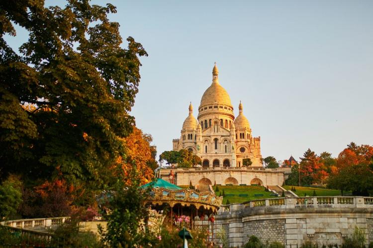 View of Sacré-Cœur Basilica at sunset, with a vintage carousel in the foreground, surrounded by autumn trees in Montmartre, Paris.