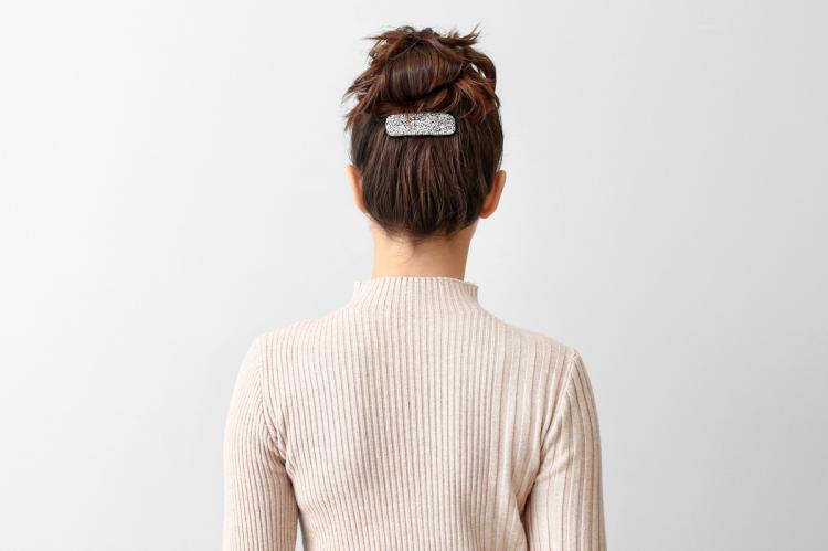 Back view of a woman with dark hair in a slicked-back bun, secured with a silver hair clip, wearing a beige ribbed sweater.