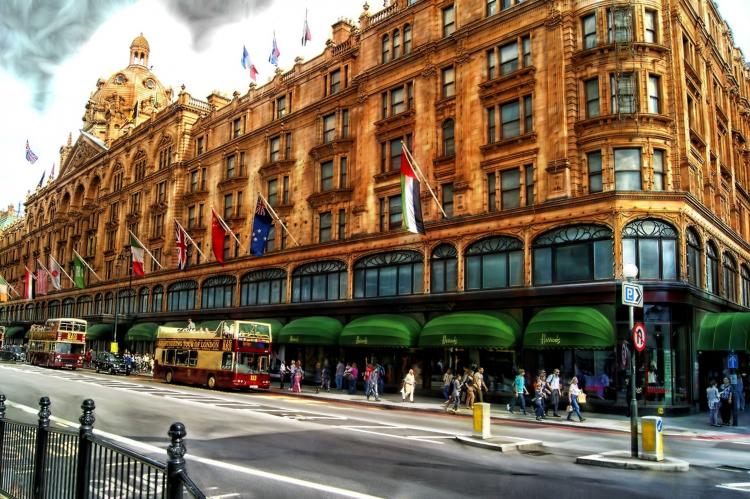Exterior of Harrods in London, showcasing its iconic architecture with ornate details, green awnings, and flags of various countries, with pedestrians and double-decker buses passing by.