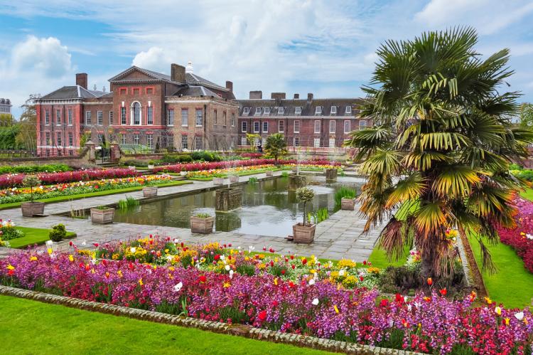 Kensington Palace in London, with its red-brick façade, surrounded by manicured gardens, vibrant flowerbeds, and a central pathway, set under a sunny sky with visitors enjoying the tranquil setting.