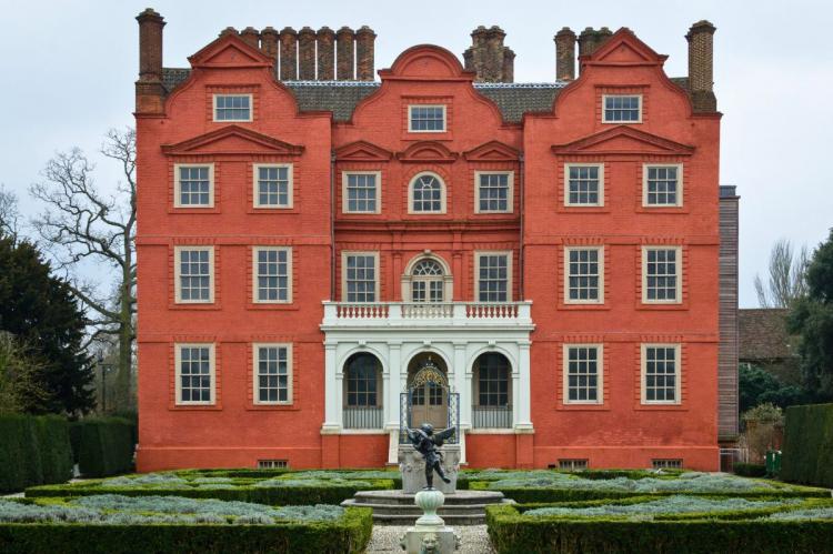 Kew Palace with its red-brick façade, symmetrical windows, and formal garden featuring neatly trimmed hedges and a fountain with a bronze statue in the foreground.