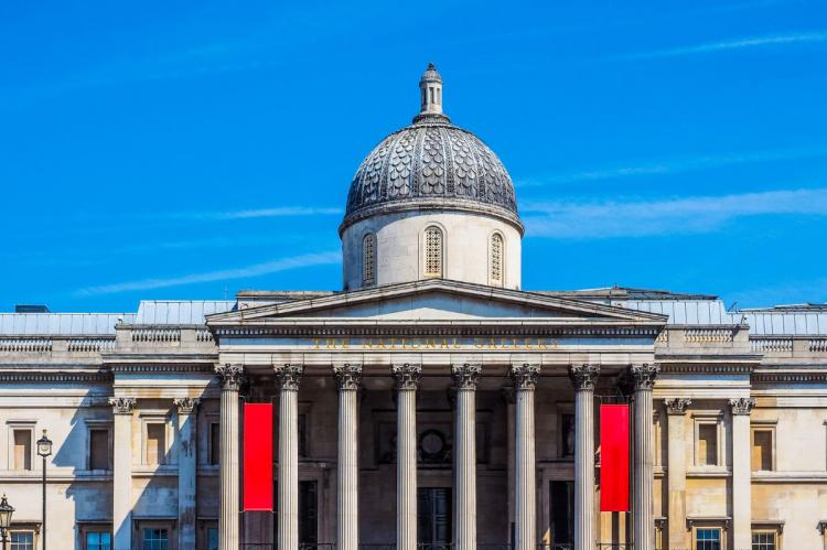 Exterior view of the National Gallery in London, featuring a neoclassical dome, Corinthian columns, and red banners, set against a bright blue sky.