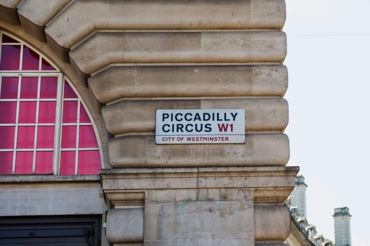 Street sign for Piccadilly Circus W1, City of Westminster, mounted on a beige stone building corner with pink-paned windows and chimney tops in the background.