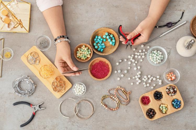 Hands making jewelry at a craft table with beads, pearls, pliers, chains, charms, and bracelets arranged in bowls and trays.