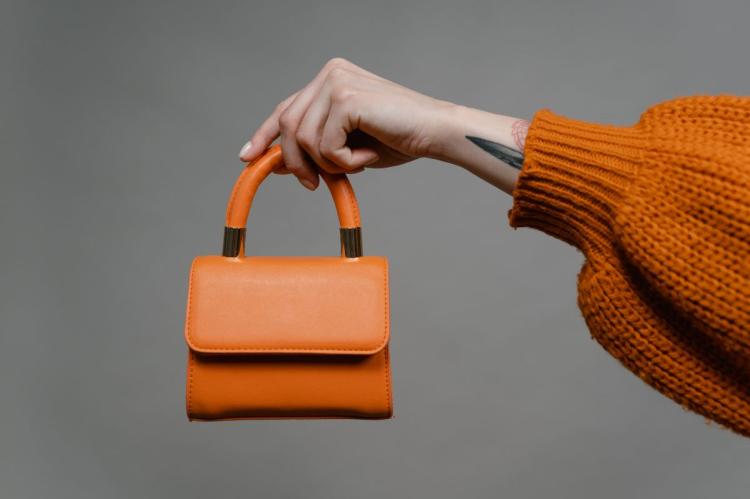 Person in an orange sweater holding a structured orange leather handbag by its top handle.