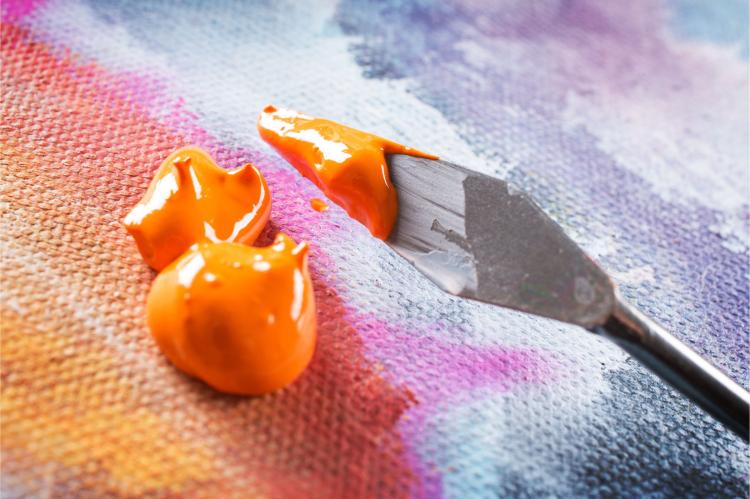 Palette knife spreading bright orange acrylic paint across a textured, multicolored canvas.