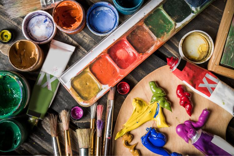 Overhead view of acrylic paints, brushes, tubes, containers, and a wooden palette covered with colorful paint.