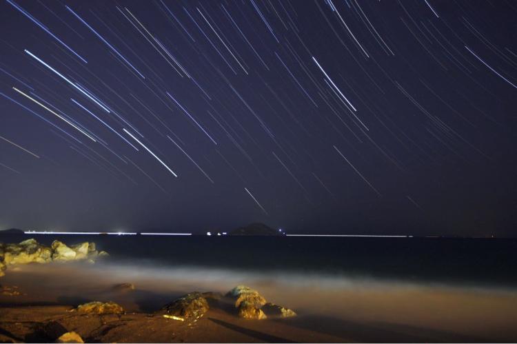Long-exposure night photo of star trails sweeping diagonally across the sky above a rocky shoreline and calm sea, with distant lights along the horizon.