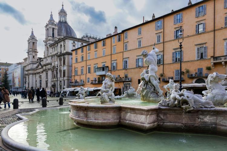Fountain with marble sculptures in Piazza Navona, surrounded by historic buildings and a domed church.