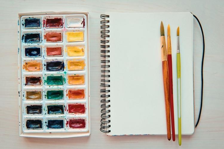 Watercolour palette beside an open blank sketchbook with three paintbrushes on a wooden table.