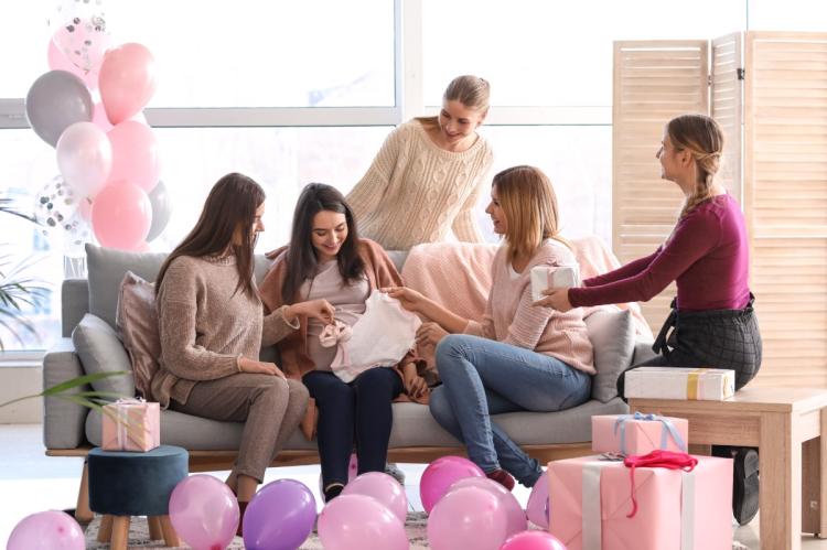 Pregnant woman opening baby clothes with friends at a baby shower surrounded by balloons and presents.