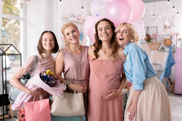Pregnant guest of honour smiling with three friends at a baby shower decorated with pink balloons.