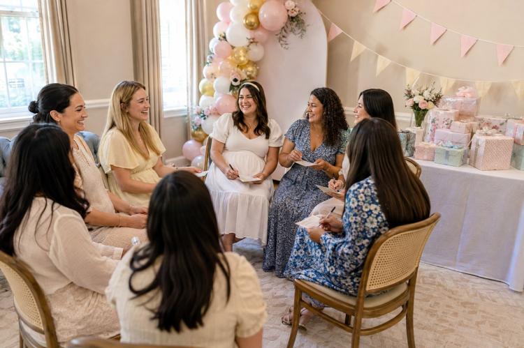 Guests seated in a circle playing a guessing game at a baby shower, with wrapped prizes on a side table.