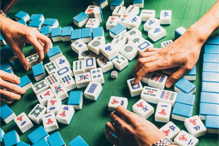 Close-up of a mahjong game in progress: many white and blue tiles marked with red, green and blue symbols scattered across a green baize, with several players' hands and a die in the centre.