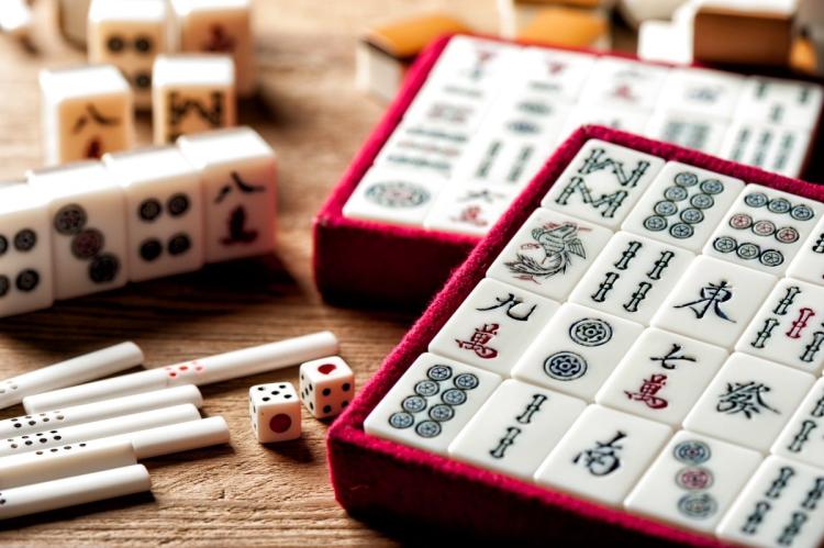 Close-up of a mahjong set: white tiles with blue, red and green markings arranged in a burgundy felt-lined tray, with two dice, a row of bone scoring sticks and loose tiles on a wooden table.