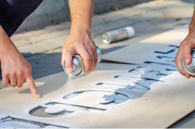Two people using spray paint and a large stencil to create a decorative design.