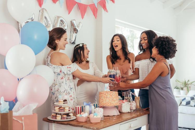 Group of women raising glasses at a baby shower party with cake, balloons and gifts.