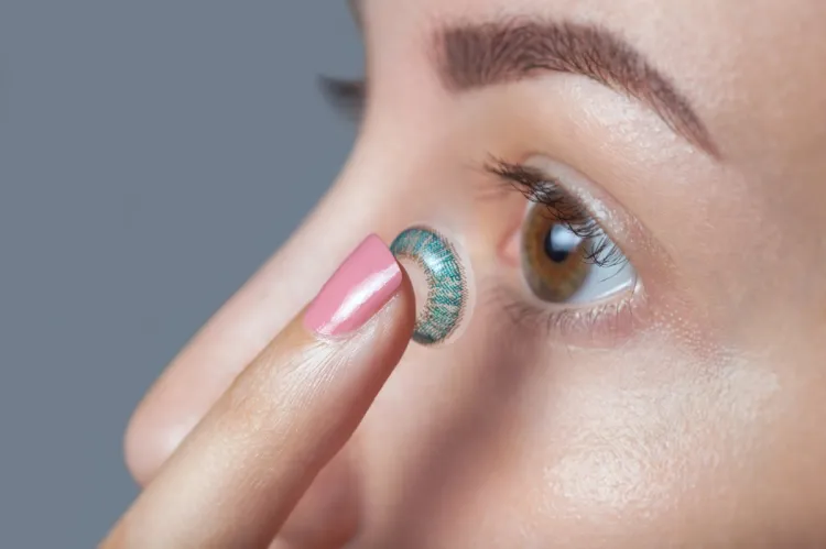 A fingertip holding a blue-green patterned contact lens up to a brown eye.