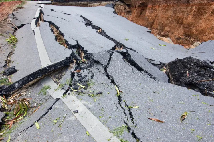 A tarmac road torn apart by an earthquake, its surface split into large slabs by deep cracks and dropped away into a red-earth ravine at one side.