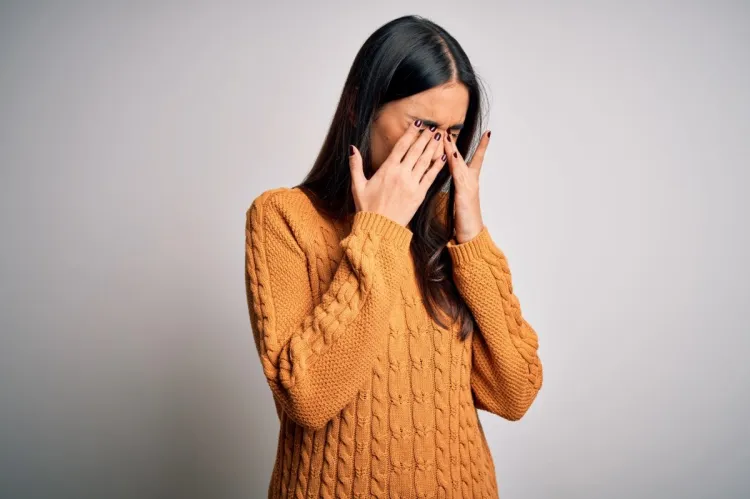 A woman in an orange jumper pressing both hands over her closed eyes.
