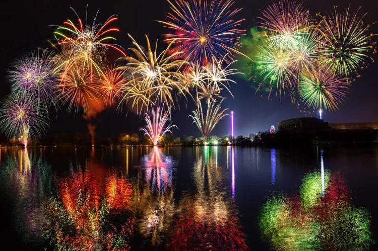 A colourful fireworks display bursting in red, gold, green, blue and pink over a calm lake at night, the bright bursts mirrored in the water with a funfair and buildings lit up on the far shore.