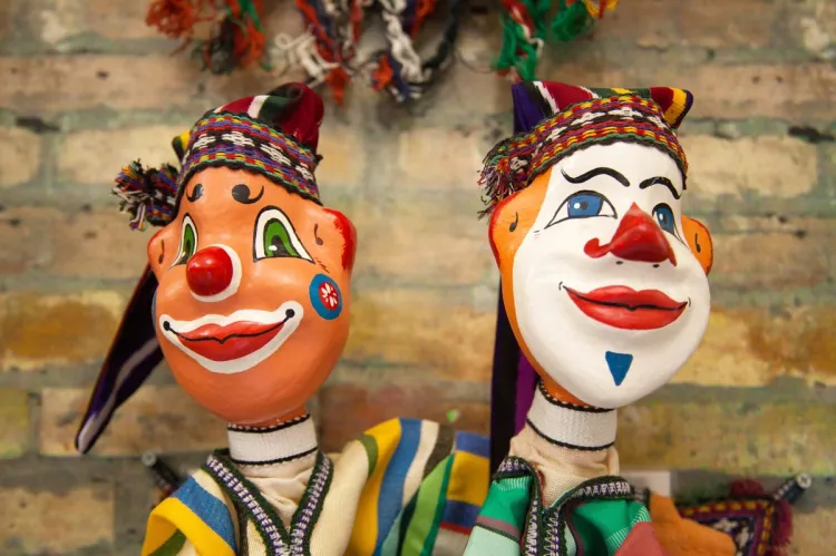 Two brightly painted papier-mâché puppet figures with clown faces, colourful woven hats and striped folk clothing, against a brick wall.